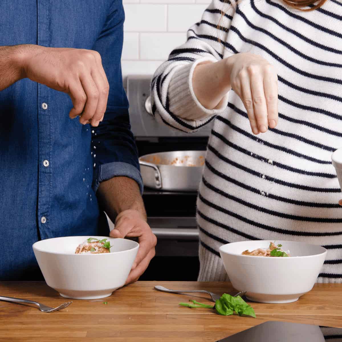 A man and woman sprinkling parmesan onto a bowl of spaghetti in their kitchen.