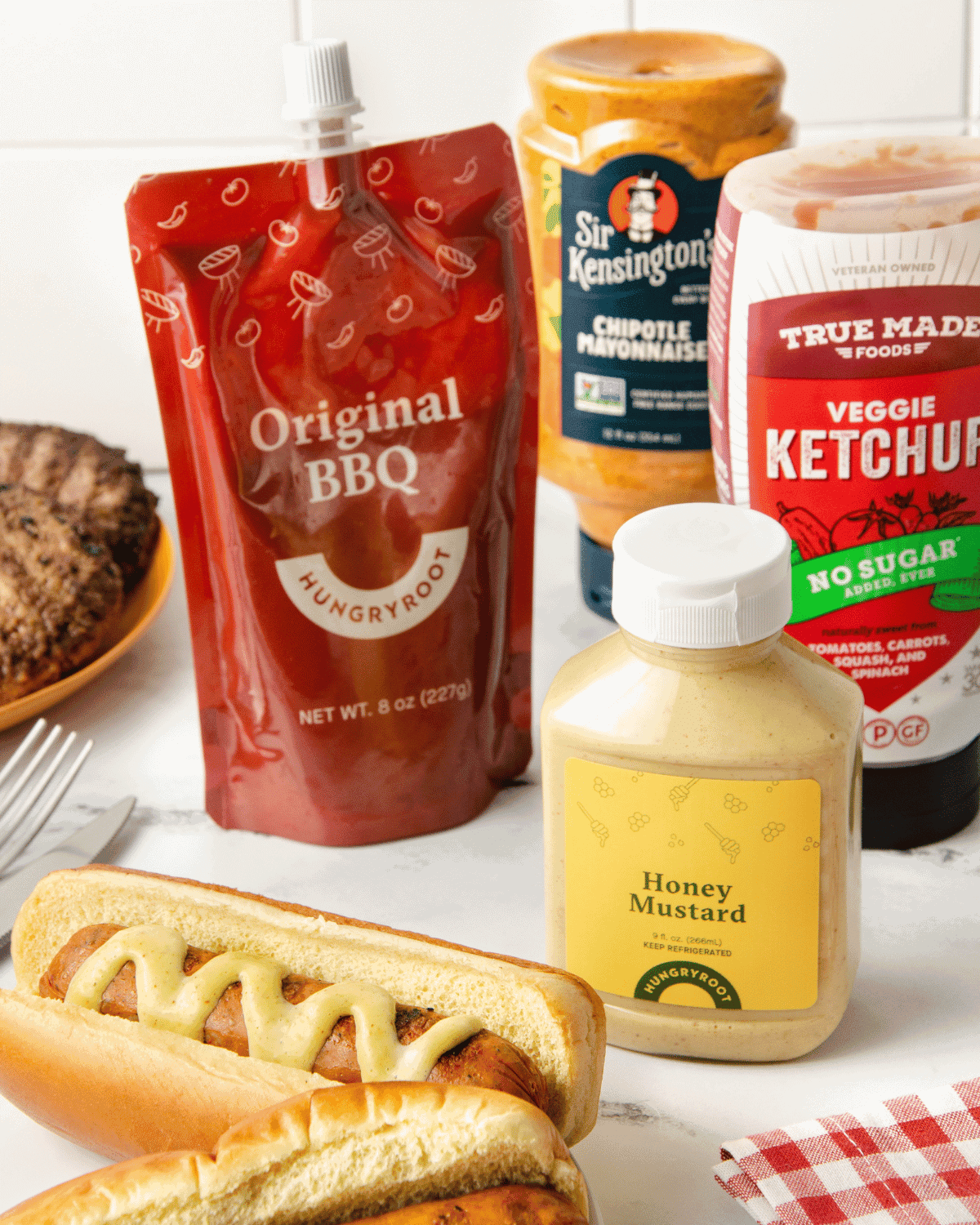 A lineup of Hungryroot's condiments behind a hot dog in a bun with mustard.