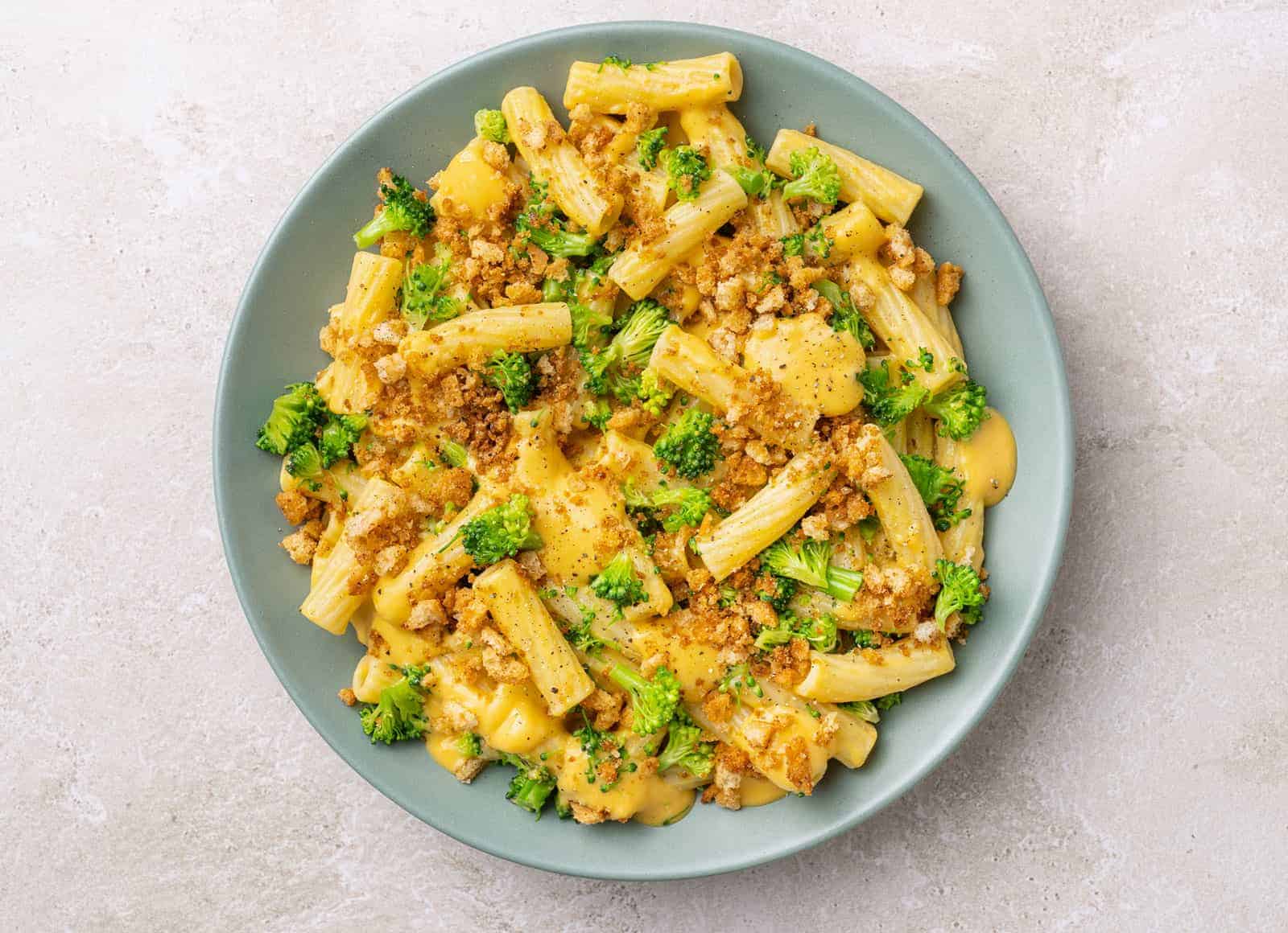 A blue plate of rigatoni tossed in a creamy cheese sauce topped with browned breadcrumbs and small chunks of broccoli.