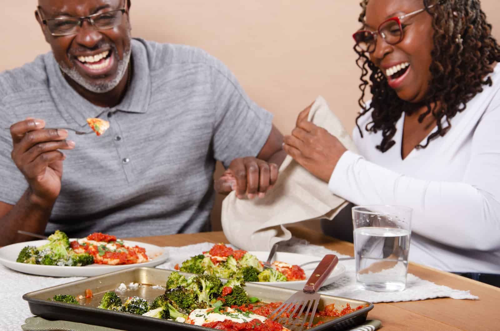 A man and woman sitting at a dinner table laughing while eating chicken parmesan.