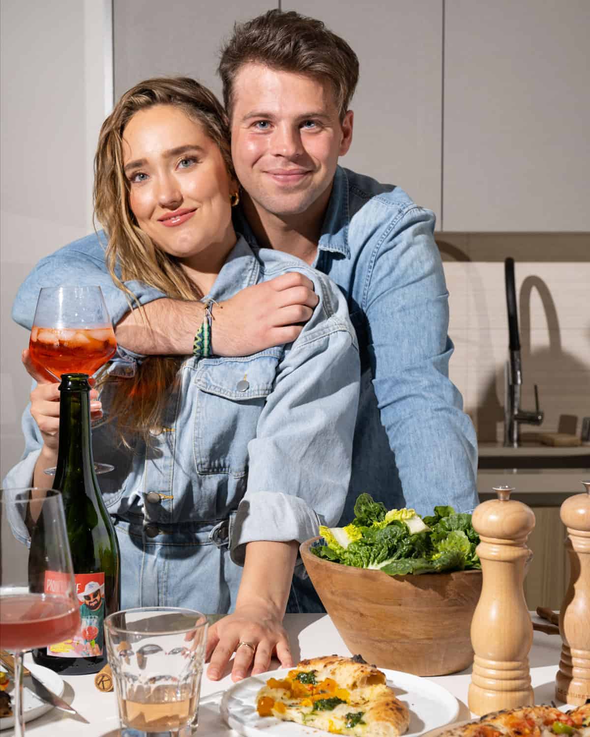 A couple standing in a kitchen holding a glass of aperol spritz in front of a whole scene of food.