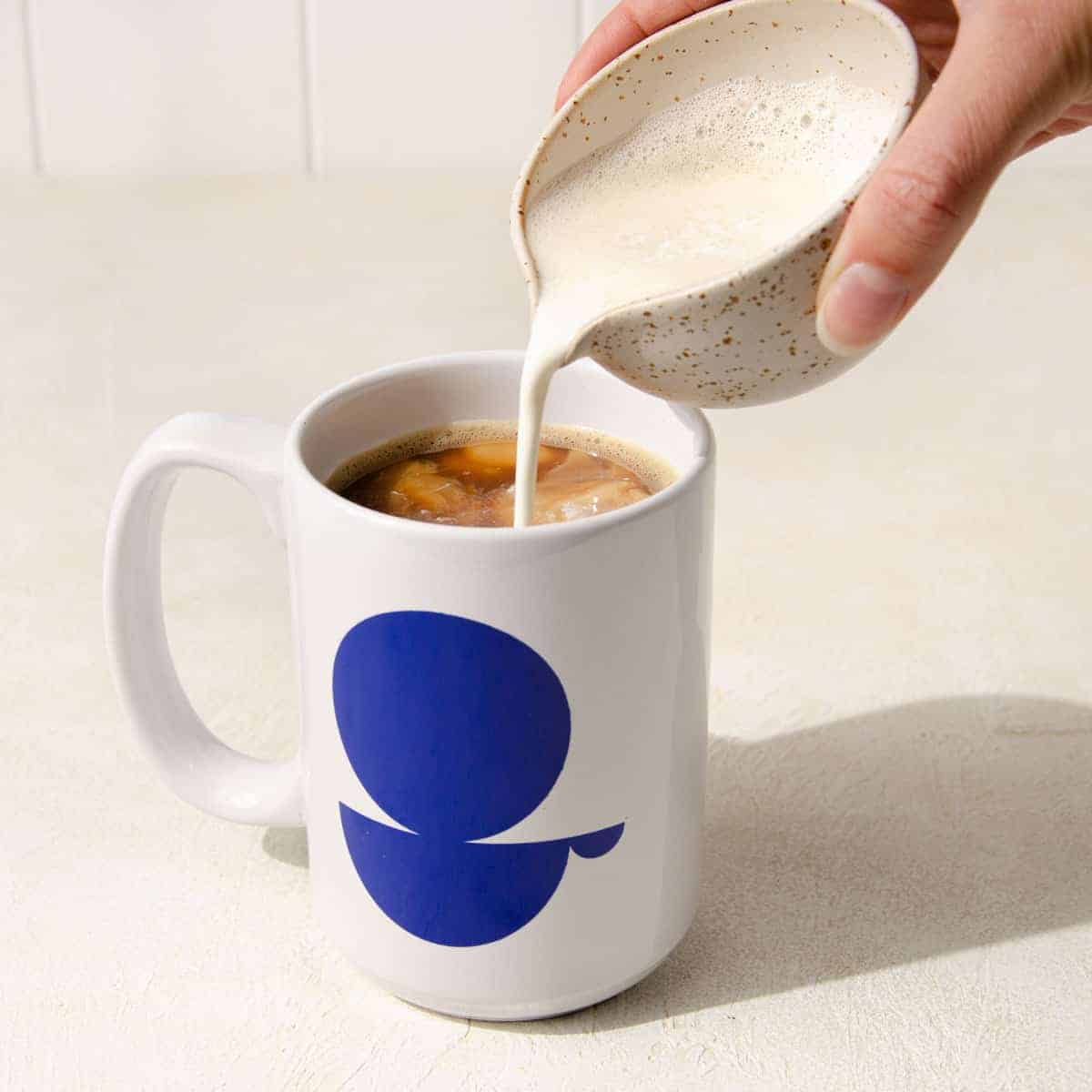 A hand pouring milk into a white mug filled with coffee.