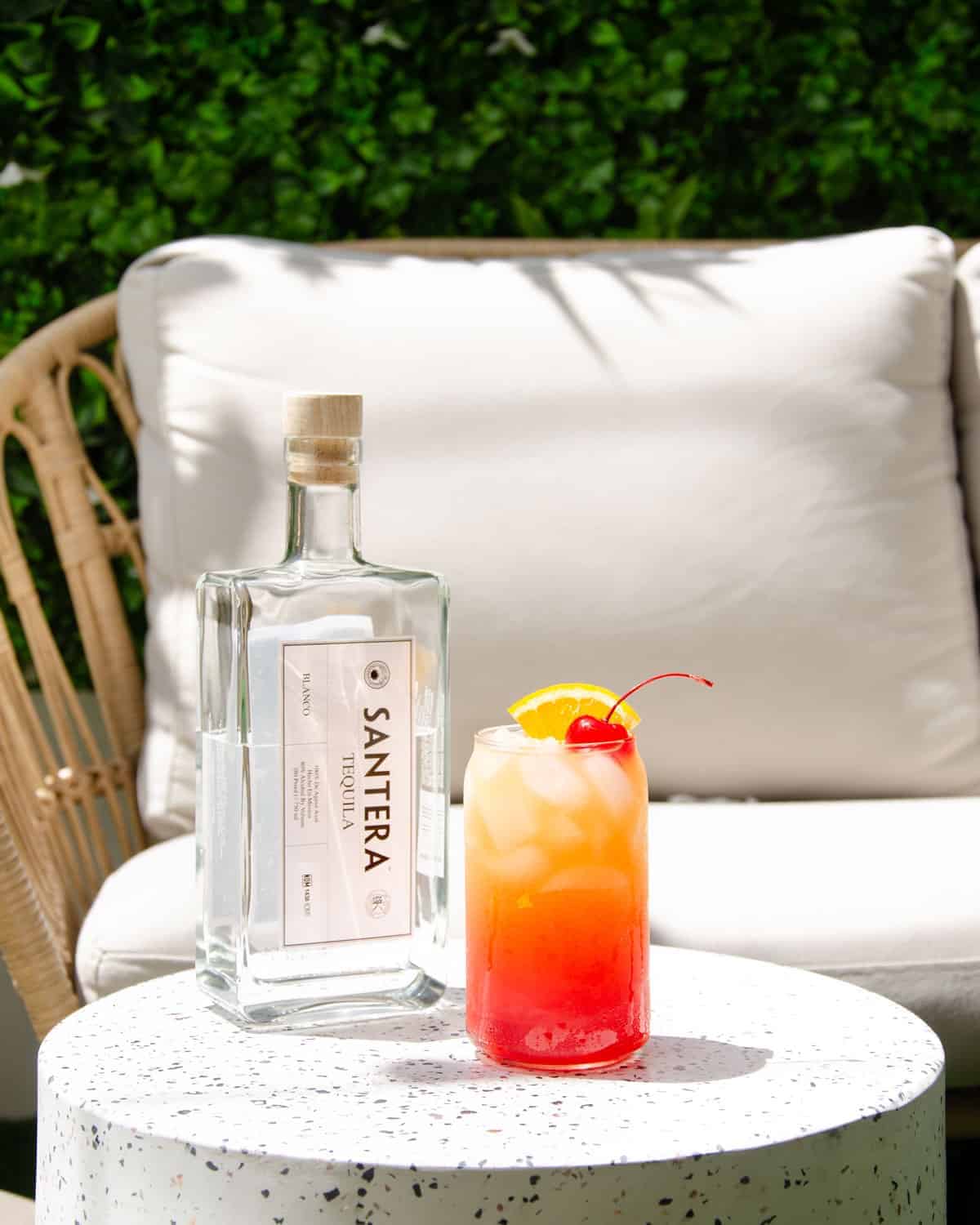 A tequila sunrise sat on a terrazzo table with a bottle of Santera tequila, in front of a patio scene.
